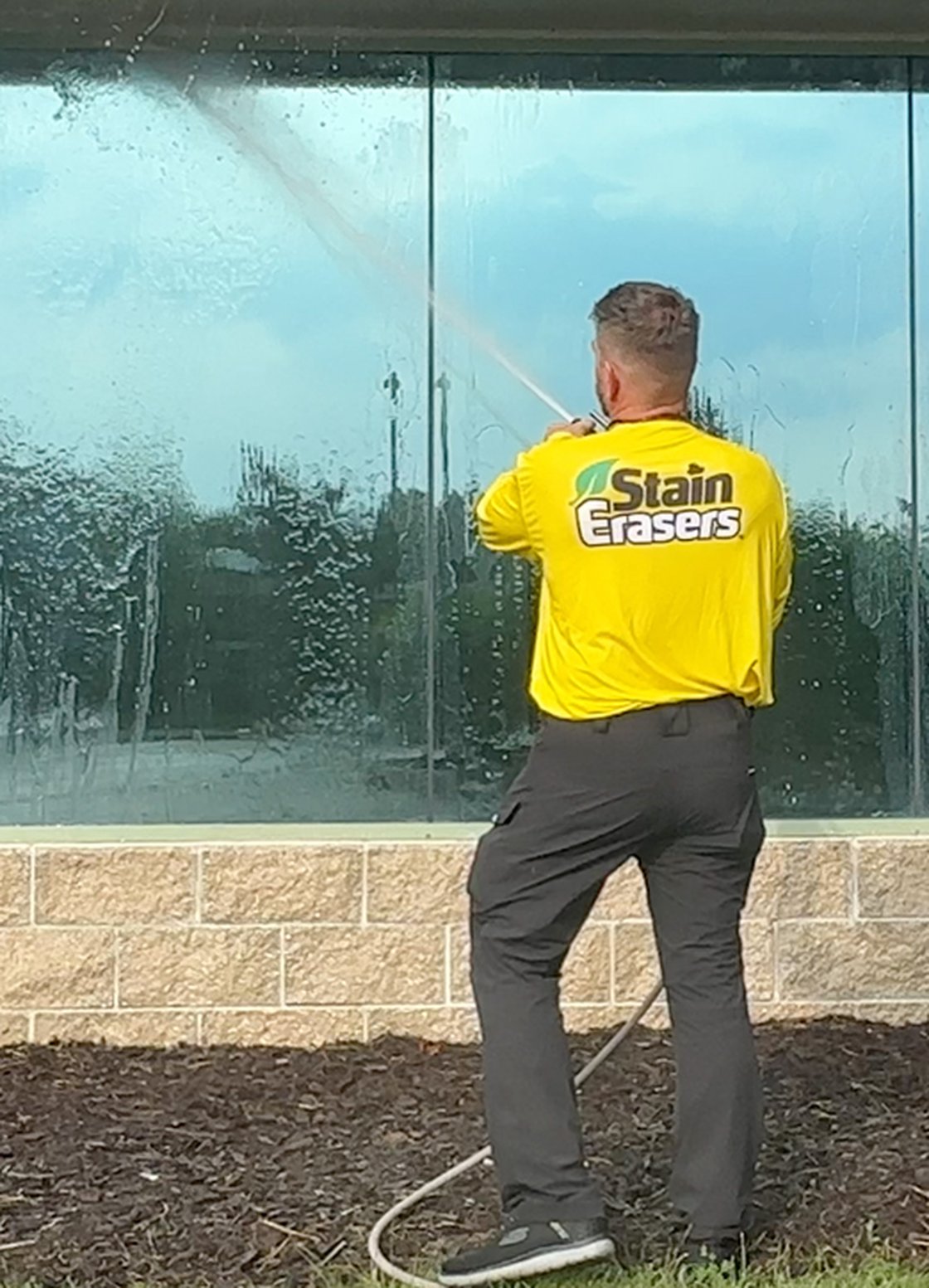 A Stain Erasers technician soft-washing a wall of storefront windows on a bright day